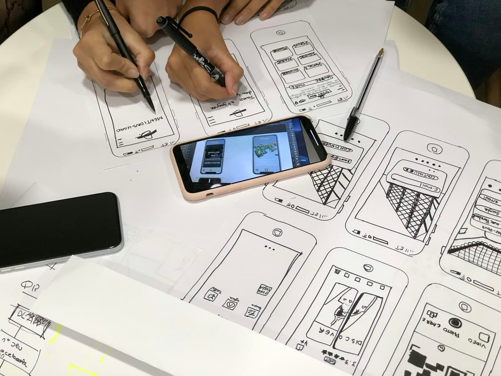 Hand-drawn mobile app wireframes on paper, with a smartphone showing a finished screen design.