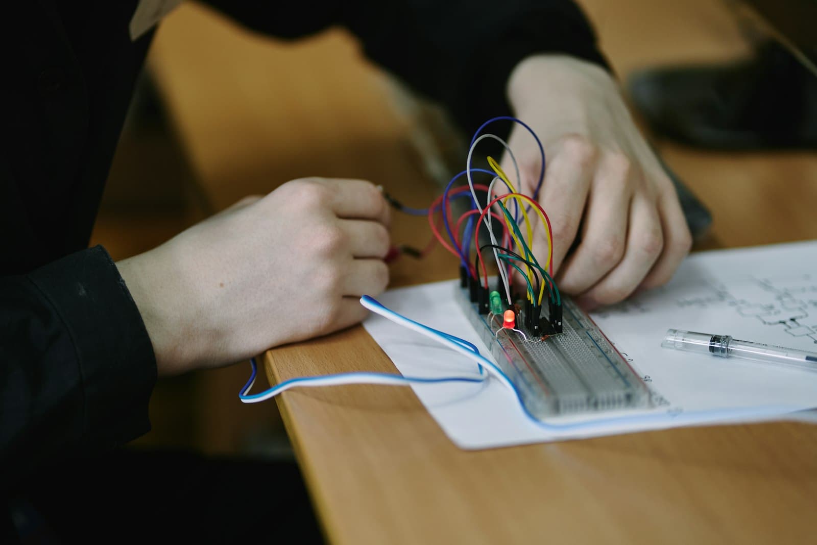 Hands connecting coloured jumper wires on a breadboard beside a hand-drawn circuit diagram.