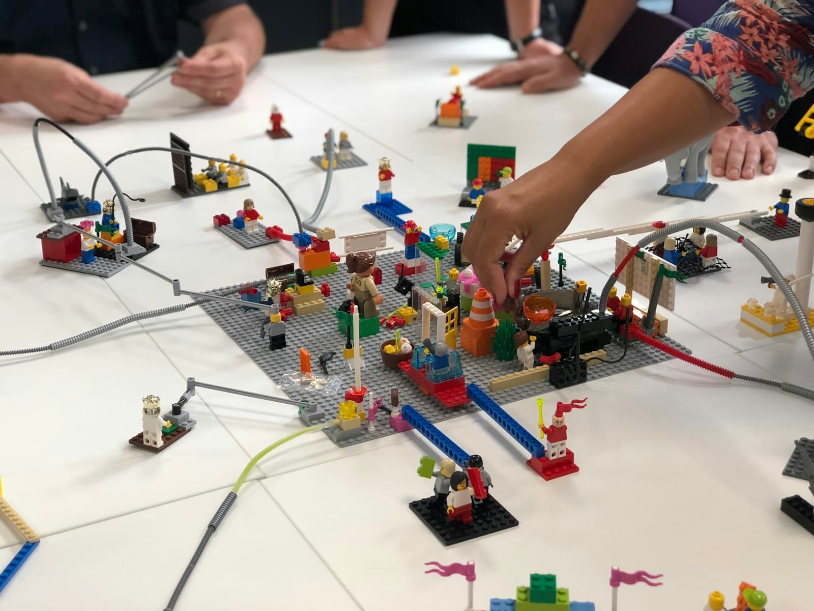 Several people's hands building a shared model town from toy bricks connected by cables on a workshop table.